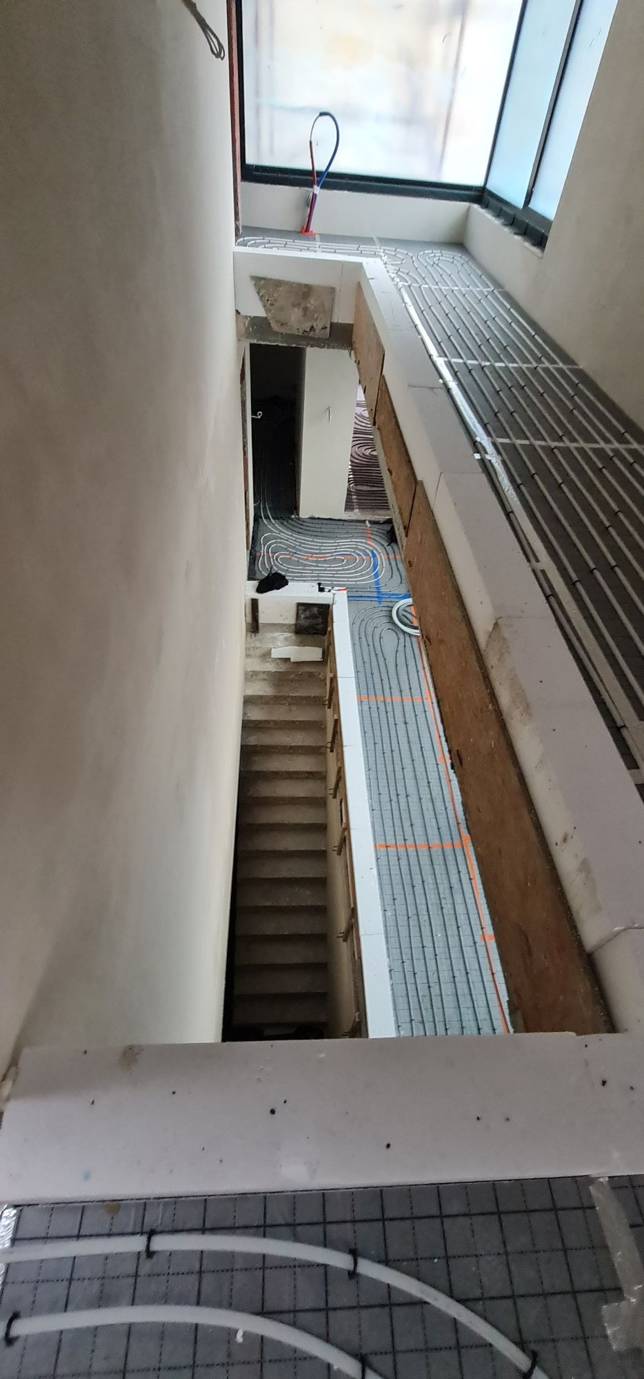 Widok z góry na instalację ogrzewania podłogowego w trakcie montażu, widoczne rury na izolacji, fragment schodów i okno dachowe.