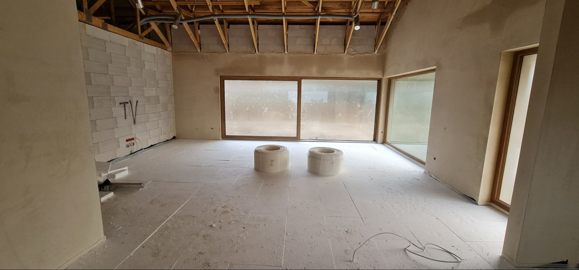 Wnętrze w trakcie wykańczania: izolacja podłogowa z rozłożonymi rolkami przewodów, ściany w surowym tynku, duże okna z drewnianą ramą i widoczne elementy instalacji grzewczej na ścianie z napisem...