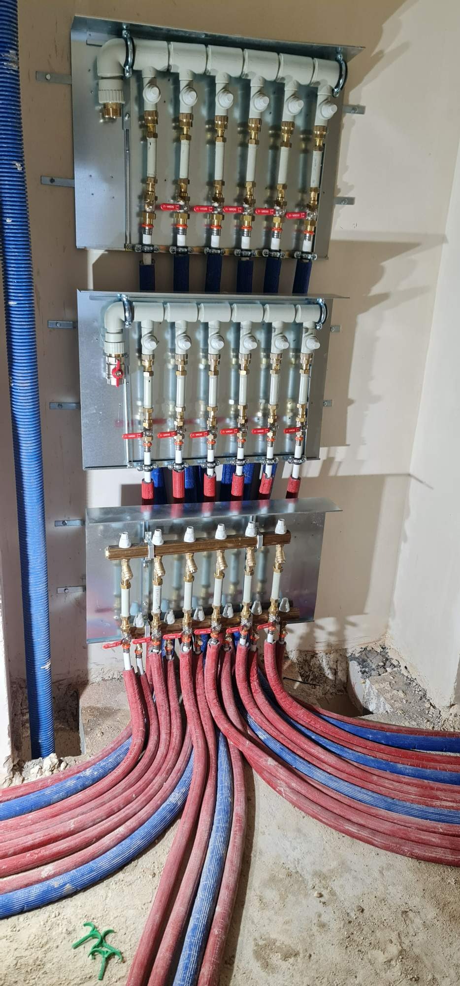 Rozdzielacz instalacji grzewczej podłączony do rur czerwonych i niebieskich, zamontowany na ścianie w surowym stanie.