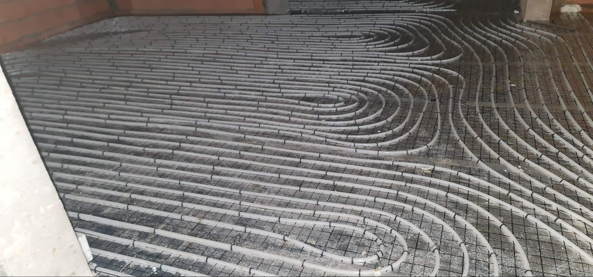 Układanie rur ogrzewania podłogowego na siatce zbrojeniowej w surowym stanie, widoczne fragmenty ścian z cegły.