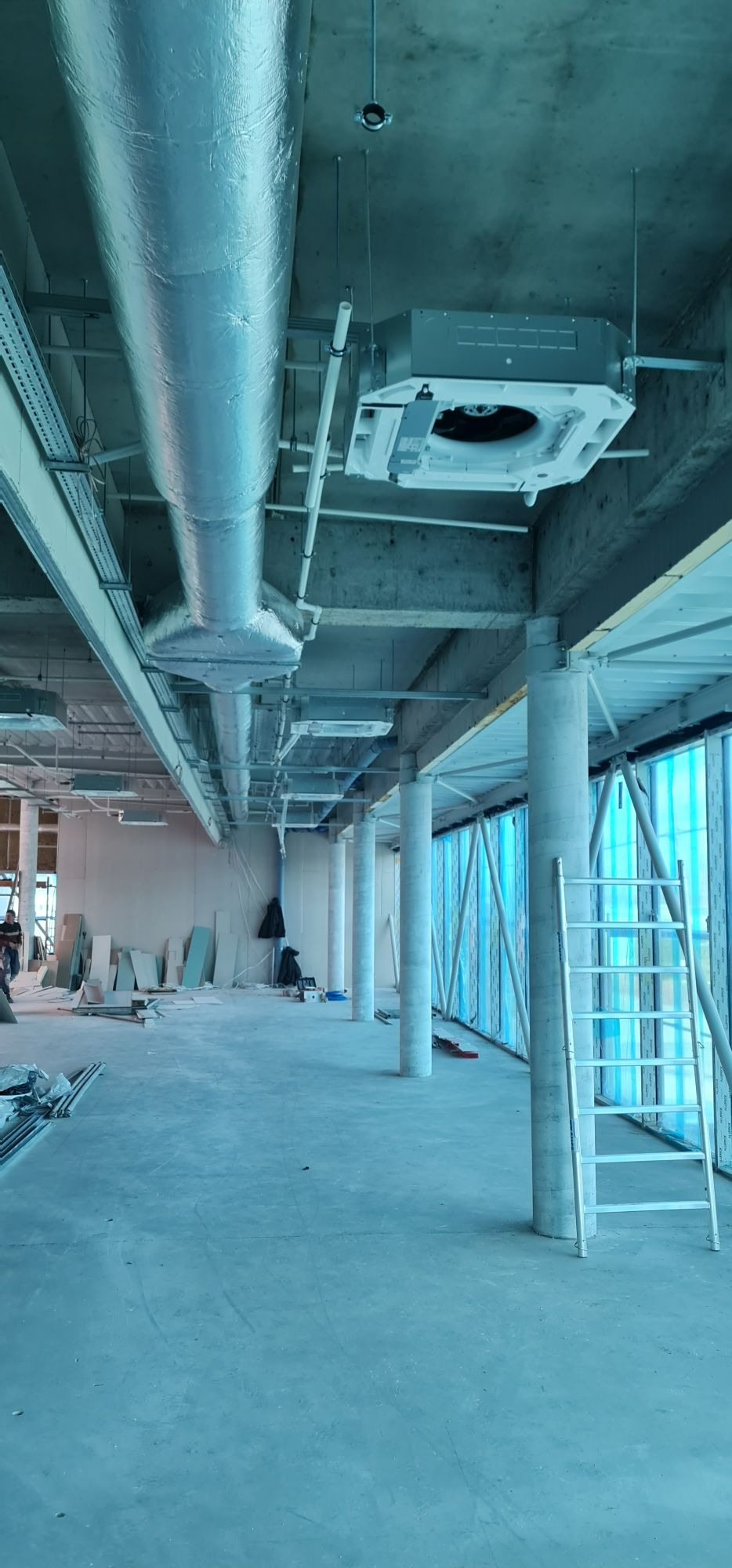 Wnętrze w trakcie instalacji systemu wentylacyjnego z widocznymi kanałami wentylacyjnymi owiniętymi folią aluminiową i jednostką centralną pod sufitem. Na podłodze widoczne materiały budowlane...