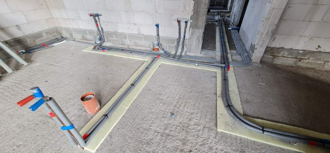 Szare rury instalacyjne ułożone w kwadraty na izolacji podłogowej w surowym pomieszczeniu z betonową posadzką i ścianami z pustaków.
