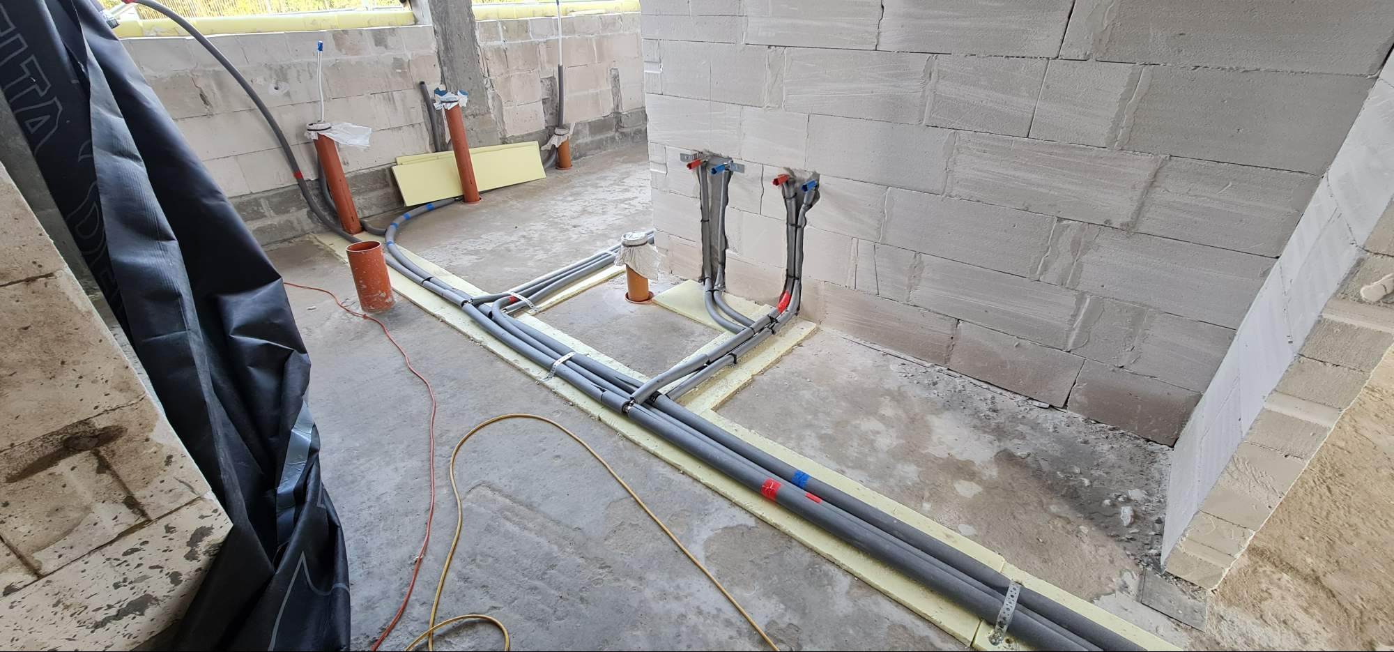 Szare rury instalacyjne prowadzone po żółtej izolacji podłogowej w surowym wnętrzu budynku z białego betonu komórkowego, widoczne oznaczenia kolorami czerwonym i niebieskim.