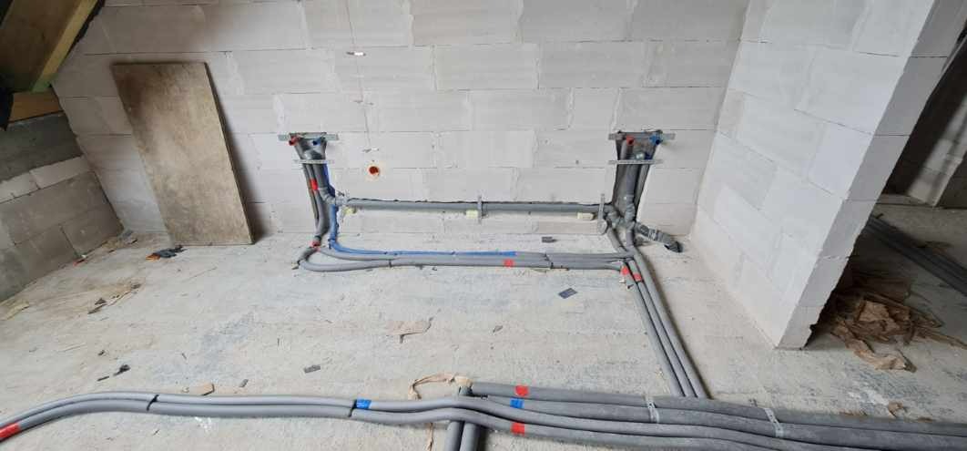 Szare rury instalacji wodnej i grzewczej prowadzone po podłodze i ścianie z białych bloczków, widoczne elementy mocujące i oznaczenia kolorami niebieskim i czerwonym.