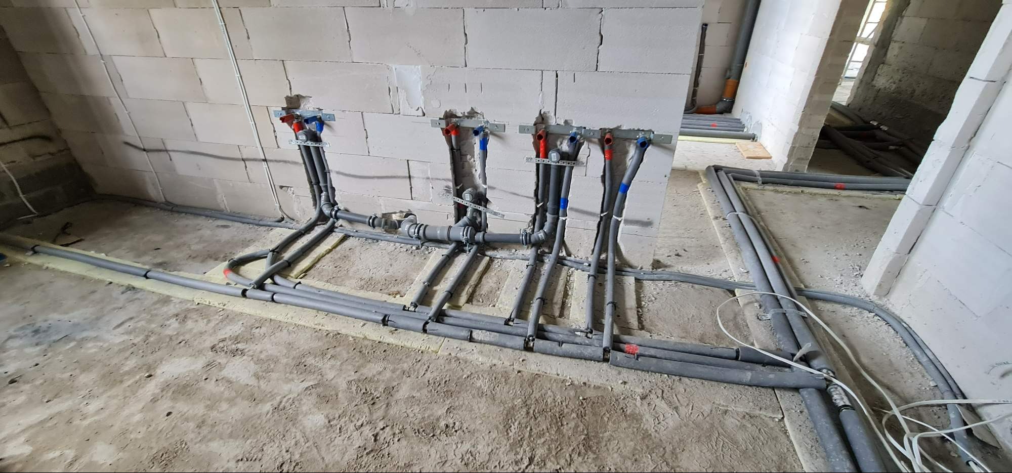 Szare rury instalacji wodnej i kanalizacyjnej w surowym stanie, zamocowane do ściany z betonu komórkowego, widoczne elementy mocujące i izolacja pod rurami.