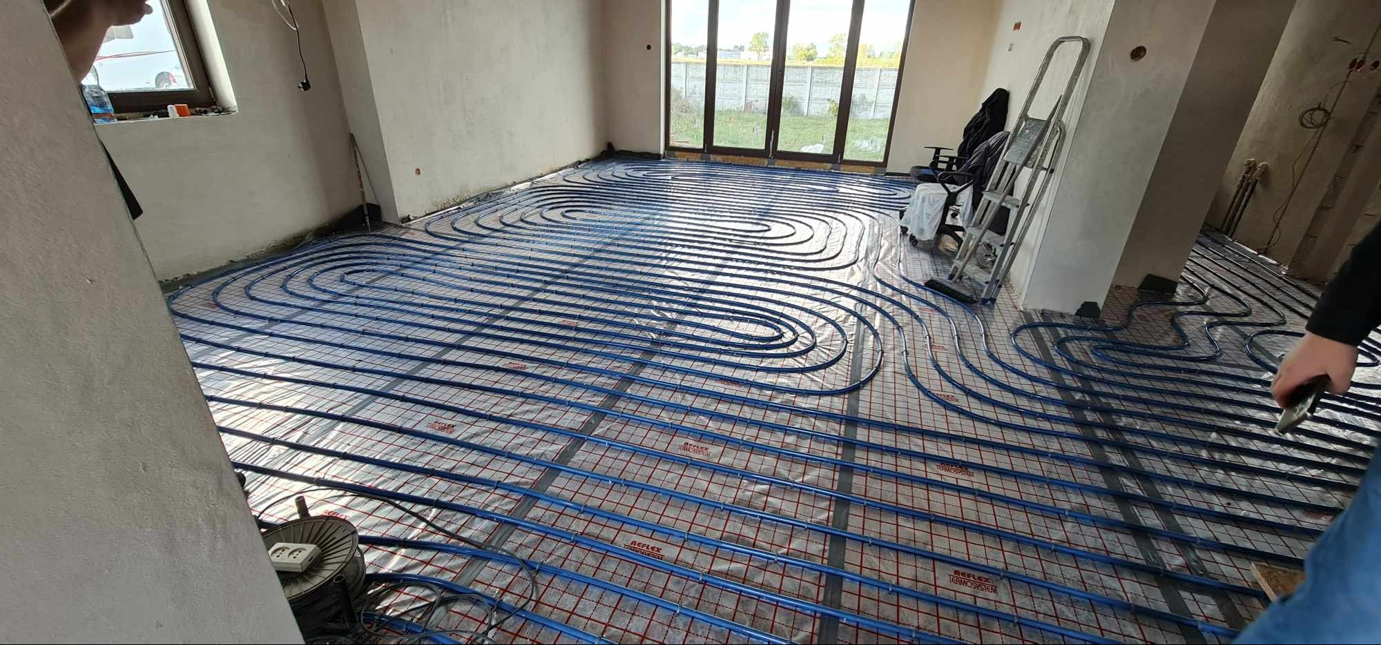 Niebieskie rury ogrzewania podłogowego ułożone spiralnie na folii refleksyjnej z czerwoną siatką w surowym wnętrzu, widoczna drabina i okno tarasowe.