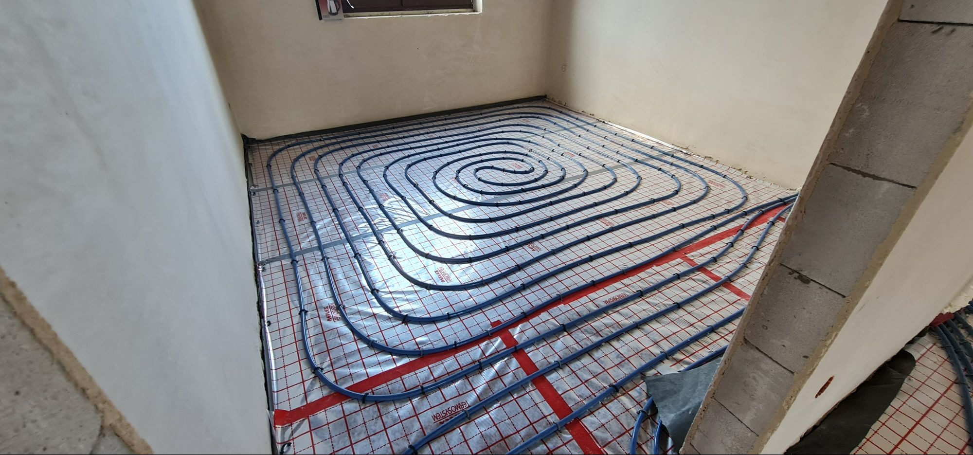 Niedokończona instalacja ogrzewania podłogowego z niebieskimi rurami ułożonymi spiralnie na srebrnej macie z czerwoną kratką w surowym wnętrzu.