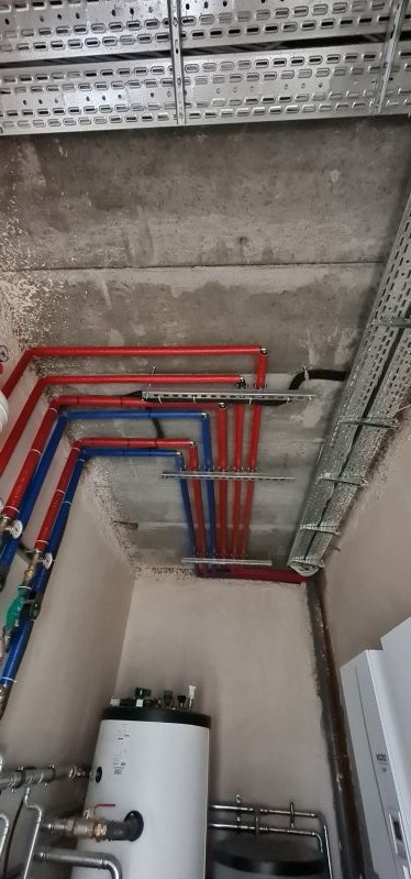 Instalacja centralnego ogrzewania z rurami w kolorze czerwonym i niebieskim oraz zasobnikiem ciepłej wody użytkowej w pomieszczeniu technicznym z surowym betonowym sufitem i korytkami kablowymi.
