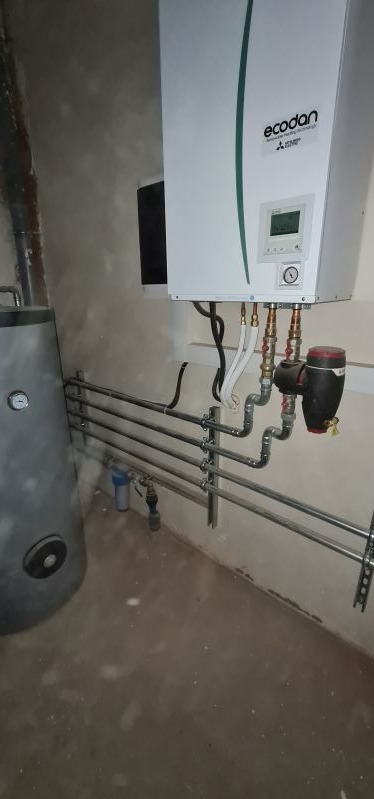 Biała jednostka grzewcza Ecodan z podłączonymi rurami miedzianymi i elementami hydraulicznymi, zamontowana na szarej ścianie w surowym pomieszczeniu z widocznym bojlerem.