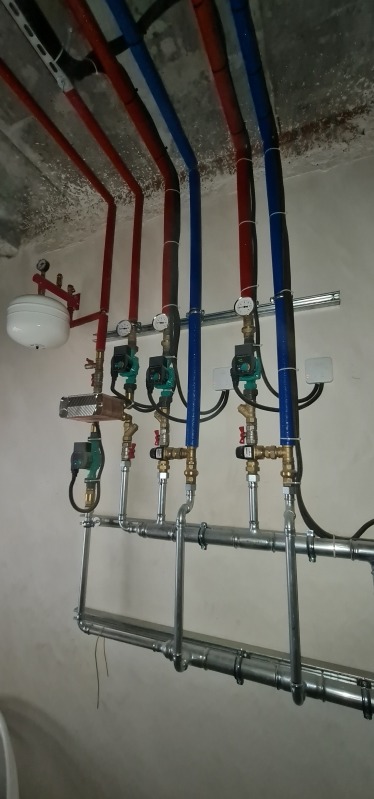Rozbudowana instalacja hydrauliczna i grzewcza z rurami w kolorach czerwonym i niebieskim, zaworami, pompami obiegowymi i manometrami, zamontowana pod sufitem w surowym stanie.