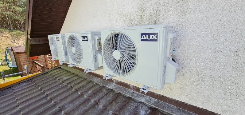 Cztery jednostki zewnętrzne klimatyzacji marki AUX zamontowane na elewacji budynku, widoczne z perspektywy dachu.