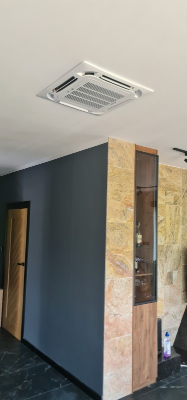 Kasetonowa jednostka klimatyzacyjna zamontowana w suficie podwieszanym, widoczna w nowoczesnym wnętrzu z ciemnoszarą ścianą i elementami wykończenia z kamienia.