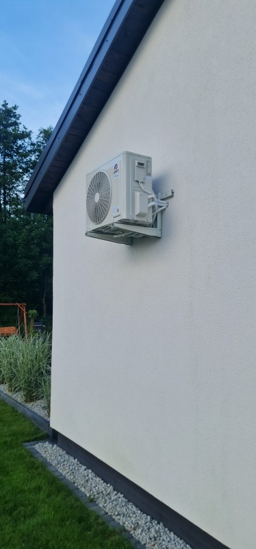 Zewnętrzna jednostka klimatyzacji marki Gree zamontowana na elewacji domu, widoczne rurki instalacyjne i wspornik montażowy, w tle trawnik i drzewa.