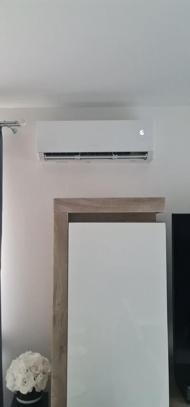 Biały klimatyzator ścienny z wyświetlaczem temperatury '16' zamontowany nad szafką w jasnym pomieszczeniu. Dekoracja z białych róż w tle.