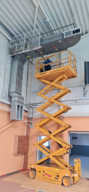 Pracownik na podnośniku nożycowym instaluje system wentylacyjny w hali, widoczne kanały wentylacyjne i kratki wentylacyjne pod sufitem.