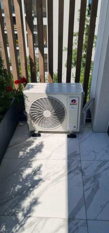 Zewnętrzna jednostka klimatyzacji marki Gree zamontowana na balkonie z marmurową posadzką, widoczne cienie drzew i kwiaty w tle.