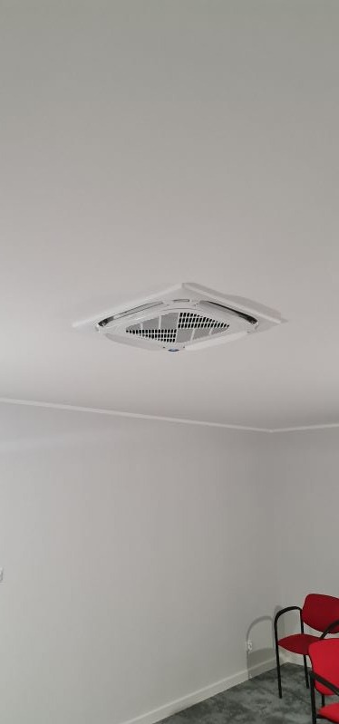Sufitowy klimatyzator kasetonowy, zamontowany w biurze z białymi ścianami i czerwonymi krzesłami w tle.