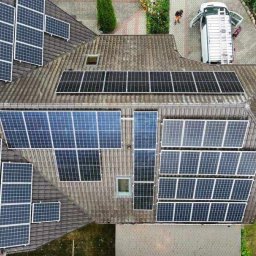 KLABAR - Widok z góry na brązowy dach domu z zainstalowanymi panelami słonecznymi o różnej wielkości i konfiguracji; biały samochód dostawczy z otwartymi drzwiami i rozłożoną drabiną obok.