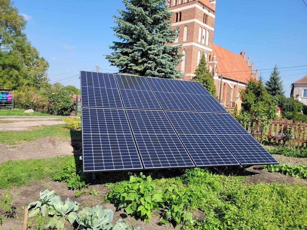 Panel słoneczny na metalowej konstrukcji w przydomowym ogródku z widokiem na ceglany kościół w tle.