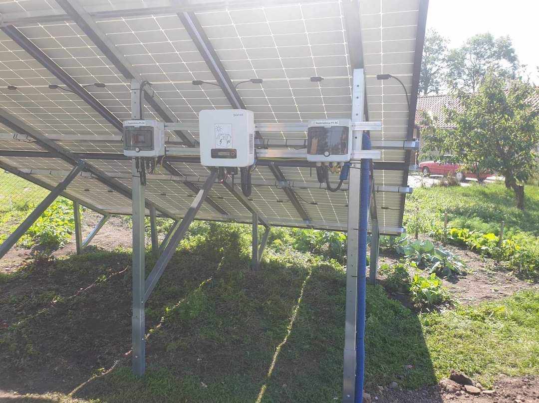 Widok od dołu na panele słoneczne zamontowane na metalowej konstrukcji, z widocznymi mikroinwerterami i niebieską rurą osłonową na kable, na tle ogrodu z warzywami i czerwonym samochodem w oddali.