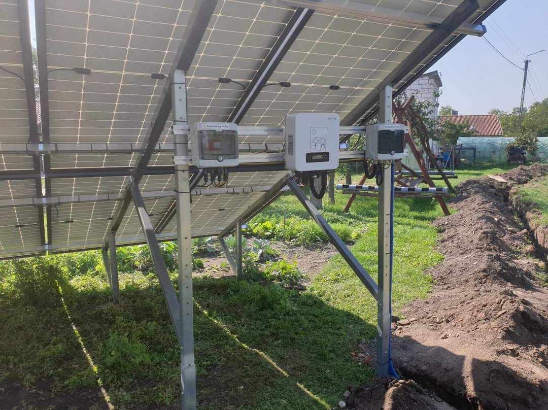 Widok od spodu na panele słoneczne z widocznymi skrzynkami przyłączeniowymi i falownikiem, na tle trawy i wykopanego rowu w ziemi.