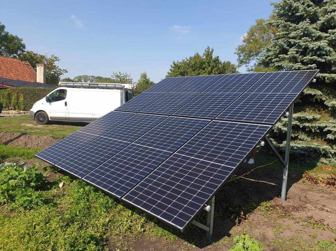 Panele fotowoltaiczne ustawione na metalowej konstrukcji na trawiastym terenie, biały samochód dostawczy w tle