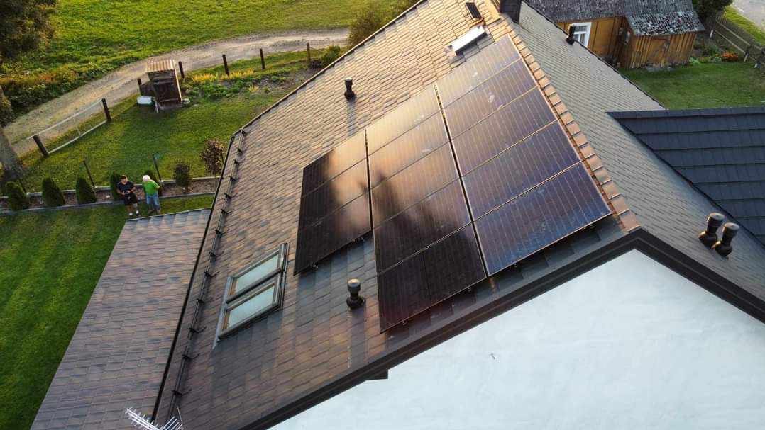 Widok z góry na dach domu z zainstalowanymi panelami słonecznymi, brązowa dachówka, widoczny fragment ogrodu z dwoma osobami.