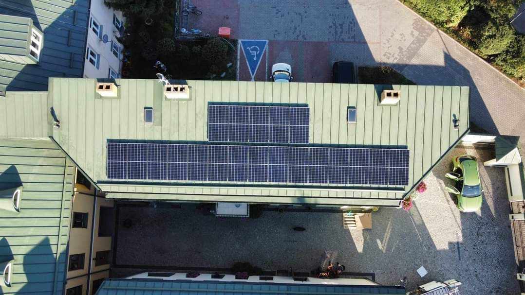 Widok z góry na budynek z panelami słonecznymi na dachu, zaparkowany samochód z otwartymi drzwiami i oznakowane miejsce parkingowe dla niepełnosprawnych.