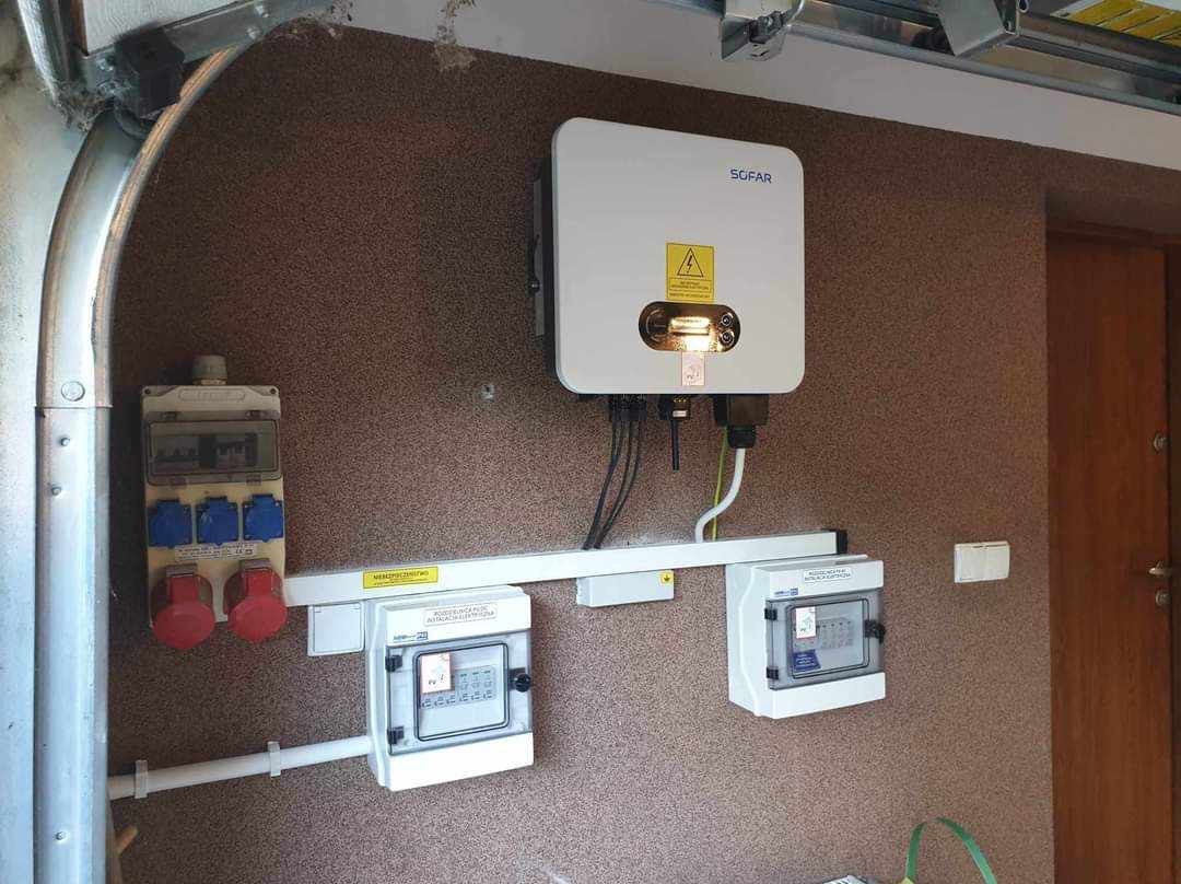 Biały falownik SOFAR z żółtą naklejką ostrzegawczą, zamontowany na ścianie garażu obok rozdzielnic elektrycznych i gniazd siłowych.