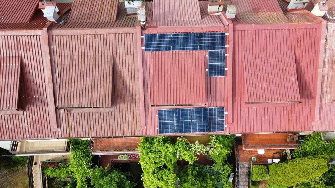 Widok z góry na czerwoną, blaszaną dachówkę z zamontowanymi panelami słonecznymi; część paneli ułożona w kształt litery L, a w tle widoczne zielone drzewa i zabudowania.