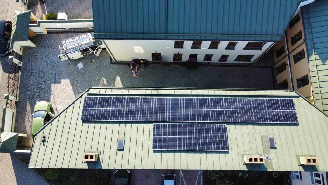 Widok z góry na budynek z panelami słonecznymi na dachu, na dziedzińcu zaparkowany biały samochód dostawczy z otwartymi drzwiami i grupa osób w pobliżu wejścia do budynku.