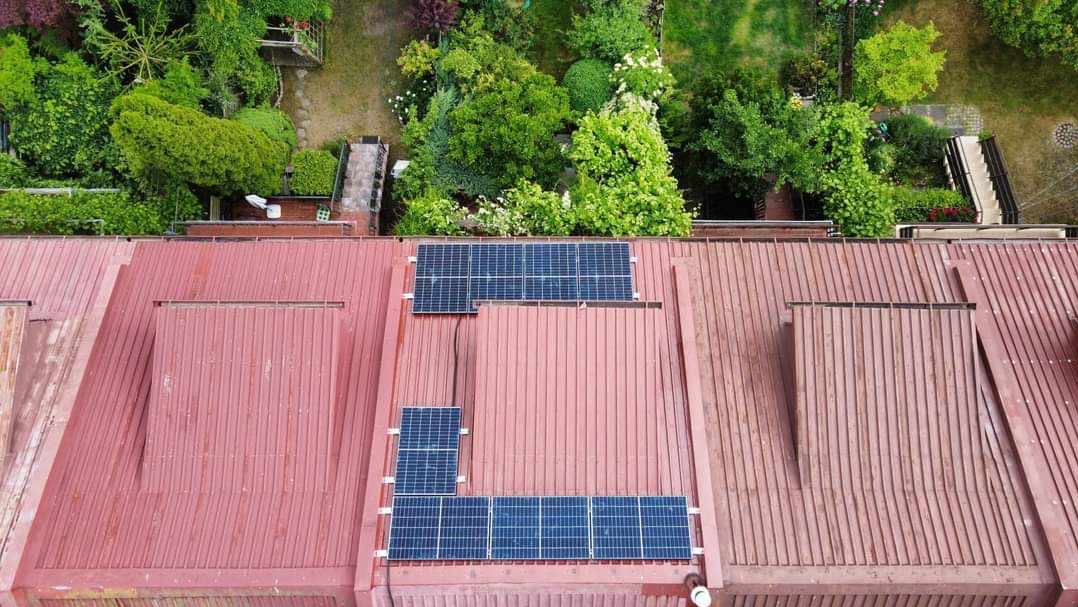 Widok z góry na panele słoneczne zamontowane na czerwonym, blaszanym dachu domu jednorodzinnego, z widocznym zielonym ogrodem w tle.