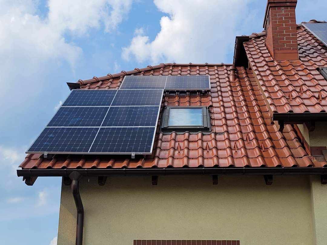 Instalacja fotowoltaiczna na dachu domu w Olsztynie z czerwoną dachówką, widoczny komin i okno dachowe, błękitne niebo w tle.