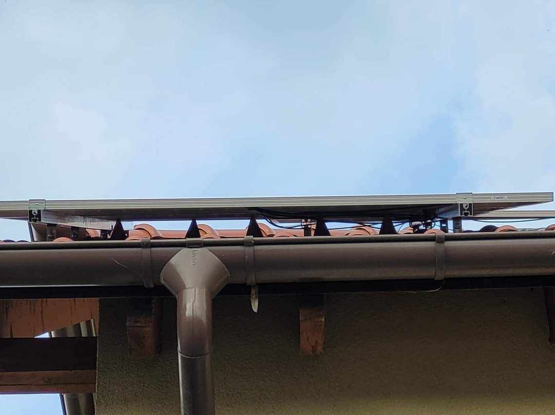 Panel słoneczny zamontowany na dachu z brązową rynną, widok z dołu na tle pochmurnego nieba.