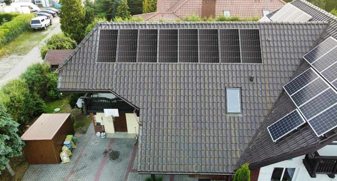 Widok z góry na dom z zainstalowanymi panelami słonecznymi na dachu z dachówki ceramicznej; część paneli w układzie poziomym, część w ukośnym; na posesji widoczny mały budynek gospodarczy i drzewa.
