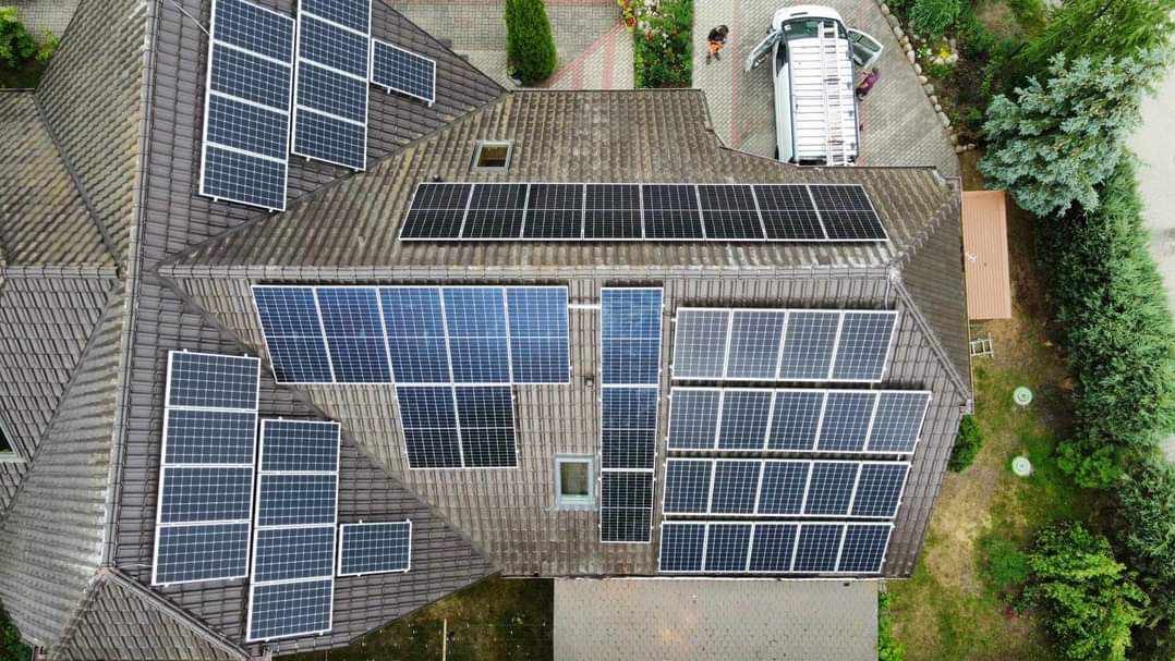 Widok z góry na brązowy dach domu z zainstalowanymi panelami słonecznymi o różnej wielkości i konfiguracji; biały samochód dostawczy z otwartymi drzwiami i rozłożoną drabiną obok.
