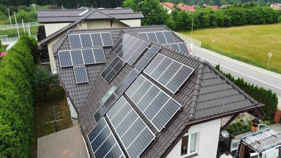 Widok z góry na dom z ciemną dachówką i panelami słonecznymi, z widoczną drogą i zielenią w tle oraz ekipą montażową przy samochodzie dostawczym