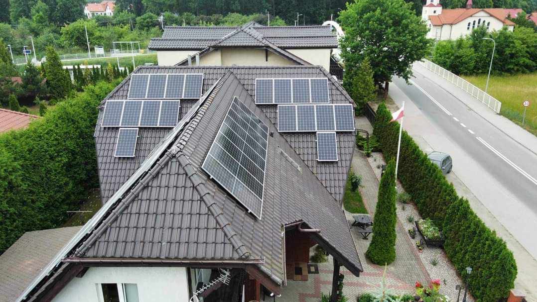 Widok z góry na dom z brązowym dachem pokrytym panelami słonecznymi; w tle budynki mieszkalne, droga i zieleń.