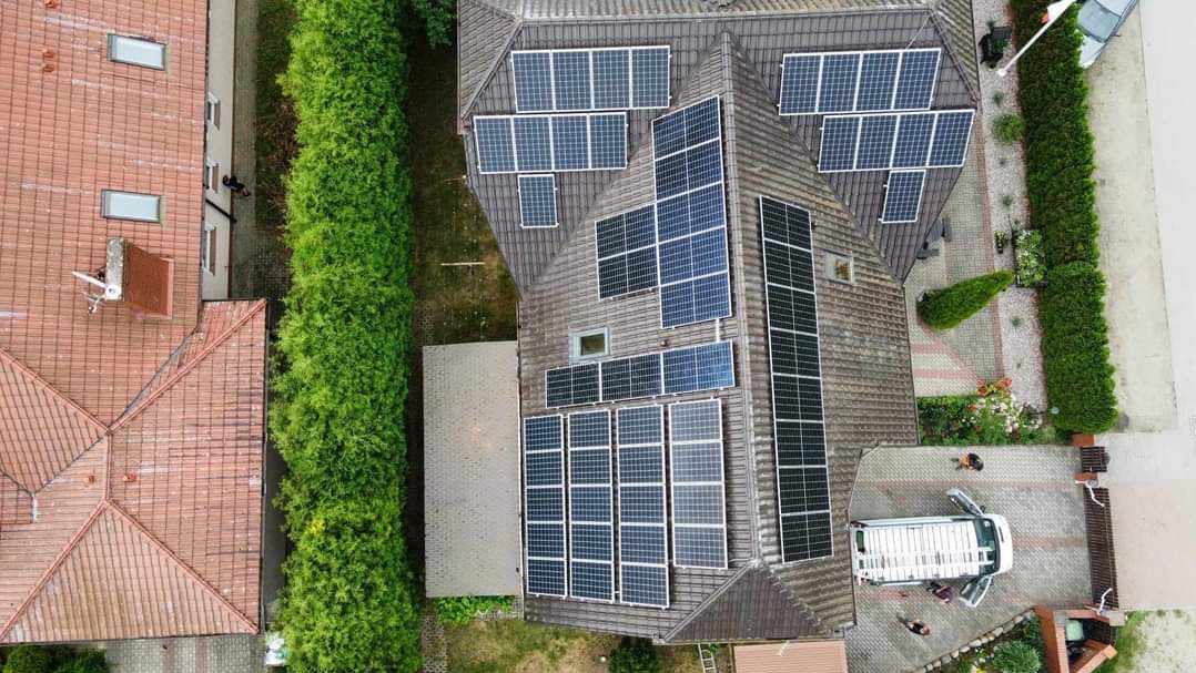 Widok z drona na dom z zamontowanymi panelami fotowoltaicznymi na dachu, obok zaparkowany biały bus z otwartymi drzwiami