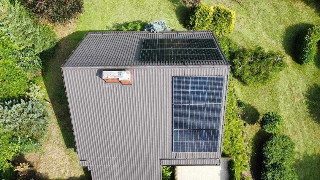 Widok z góry na dach domu z zainstalowanymi panelami słonecznymi; część paneli umieszczona w górnej części dachu, a część bliżej krawędzi; dach pokryty szarą blachą trapezową; w tle zielony trawnik...