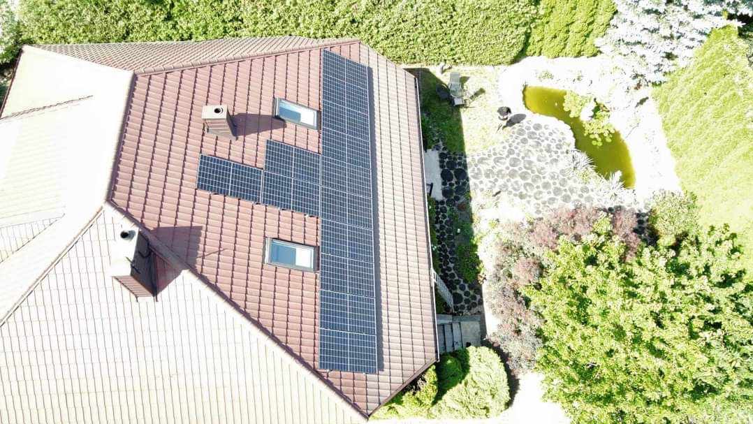 Widok z góry na dom z brązową dachówką, na której zamontowane są panele słoneczne; obok domu widoczny ogród z oczkiem wodnym.