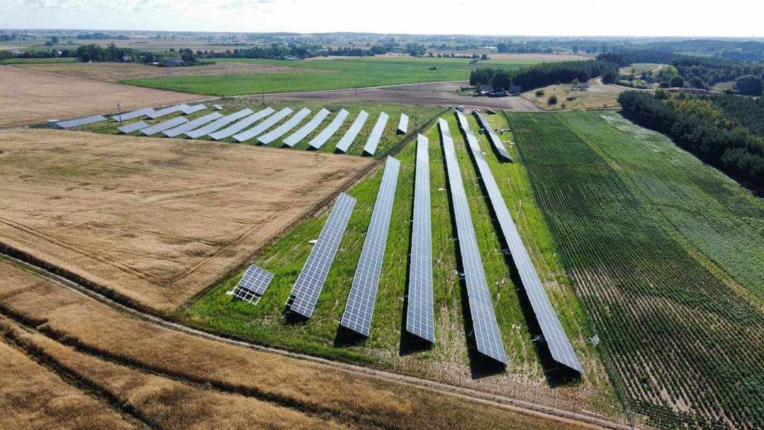 Rozległa farma paneli słonecznych na polach uprawnych, widok z lotu ptaka, zróżnicowane kolory pól i lasów w tle.