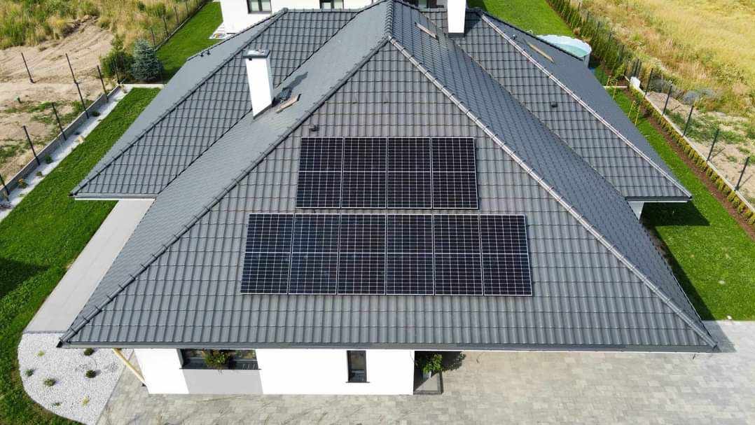 Widok z góry na dach domu jednorodzinnego z szarą dachówką ceramiczną i dwoma zestawami paneli fotowoltaicznych, otoczonego zielonym trawnikiem.