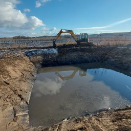 M-Kop Grzegorz Szura Prace Ziemne - Żółta koparka nad wykopem wypełnionym wodą, odbicie koparki w wodzie, w tle widoczne drzewa i błękitne niebo z chmurami.