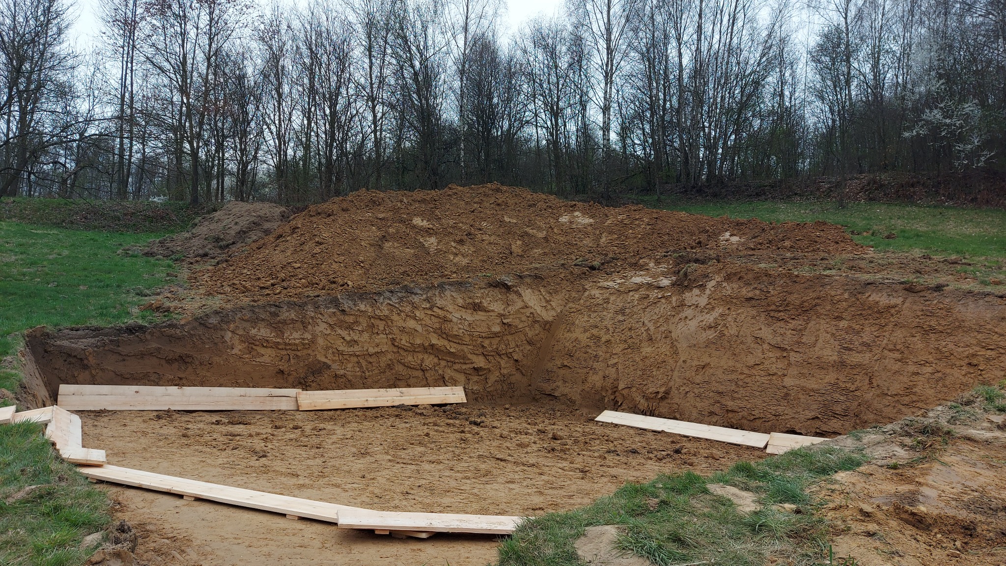 Głęboki wykop w ziemi z drewnianym szalunkiem ułożonym na dnie, pryzma ziemi obok wykopu, w tle drzewa bez liści.