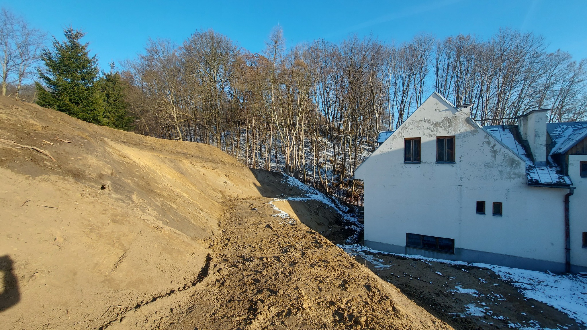 Wykopany teren przy białym budynku z widocznymi śladami śniegu i drzewami w tle, ukazujący przygotowania do prac budowlanych.