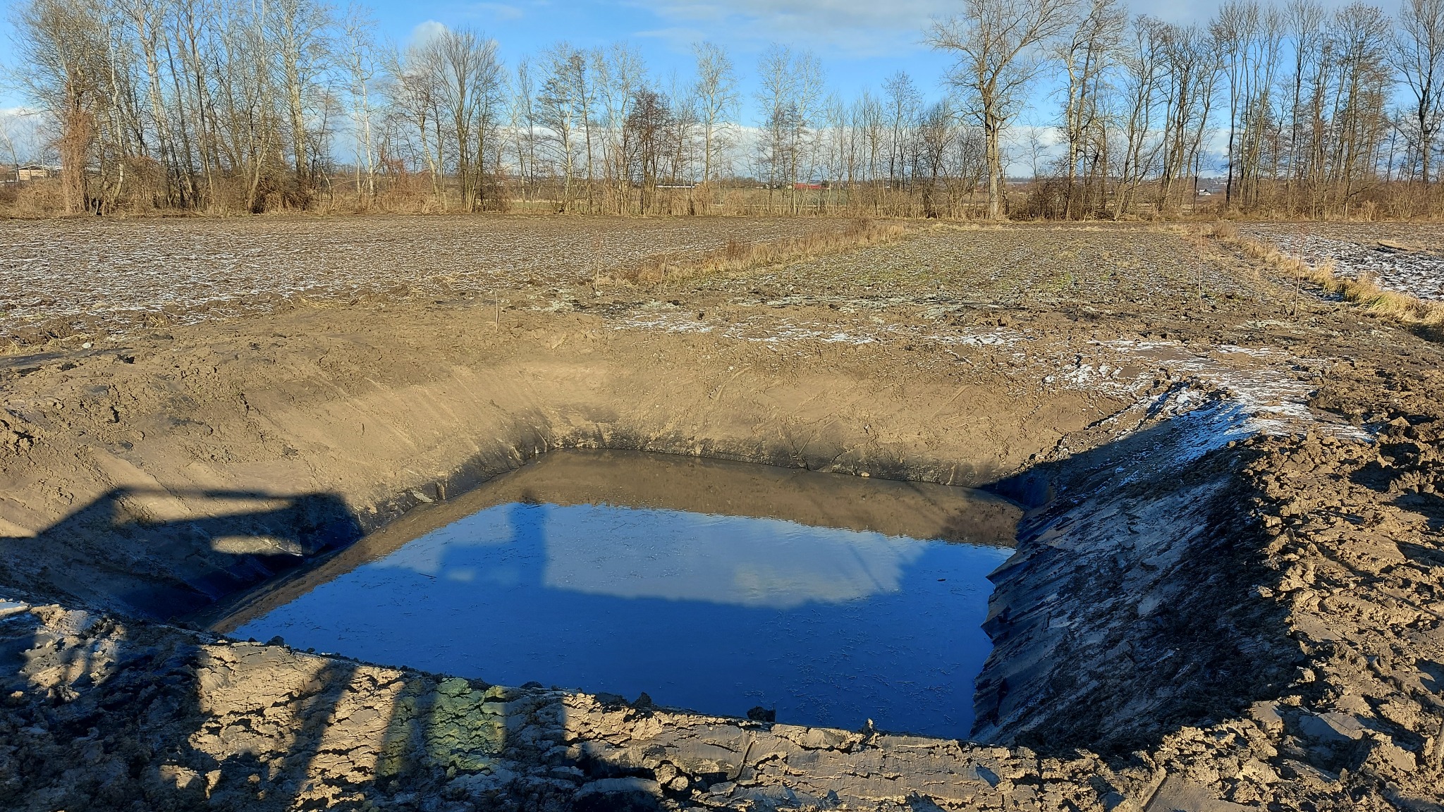Wykop pod szambo wypełniony wodą, widoczne odbicie nieba, otoczony ziemią z resztkami śniegu, w tle pole i rząd drzew bez liści.
