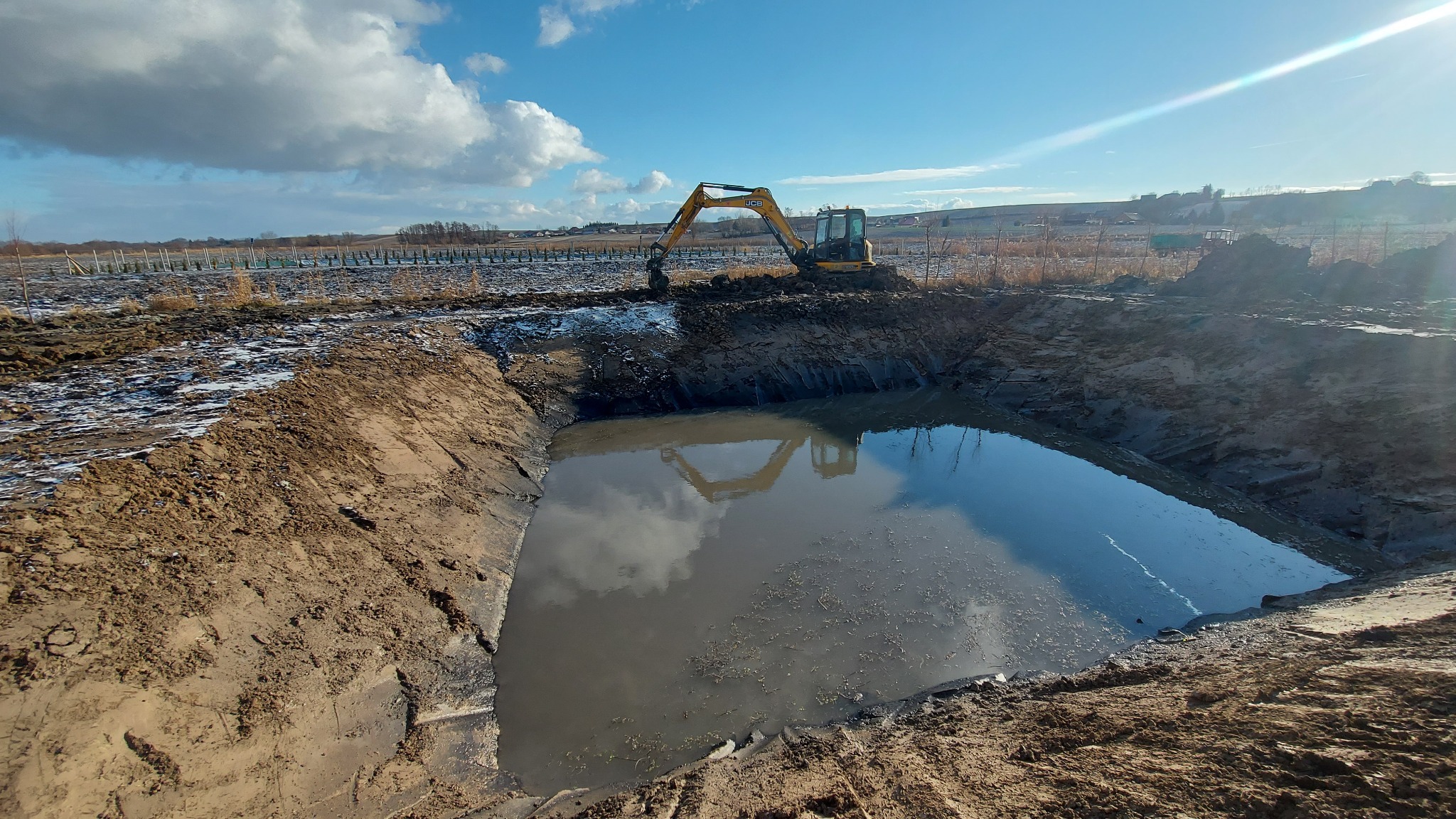 Żółta koparka nad wykopem wypełnionym wodą, odbicie koparki w wodzie, w tle widoczne drzewa i błękitne niebo z chmurami.