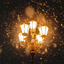 Oświetlona, ozdobna latarnia uliczna w czasie intensywnych opadów śniegu, widoczne płatki śniegu rozświetlone ciepłym światłem lamp.