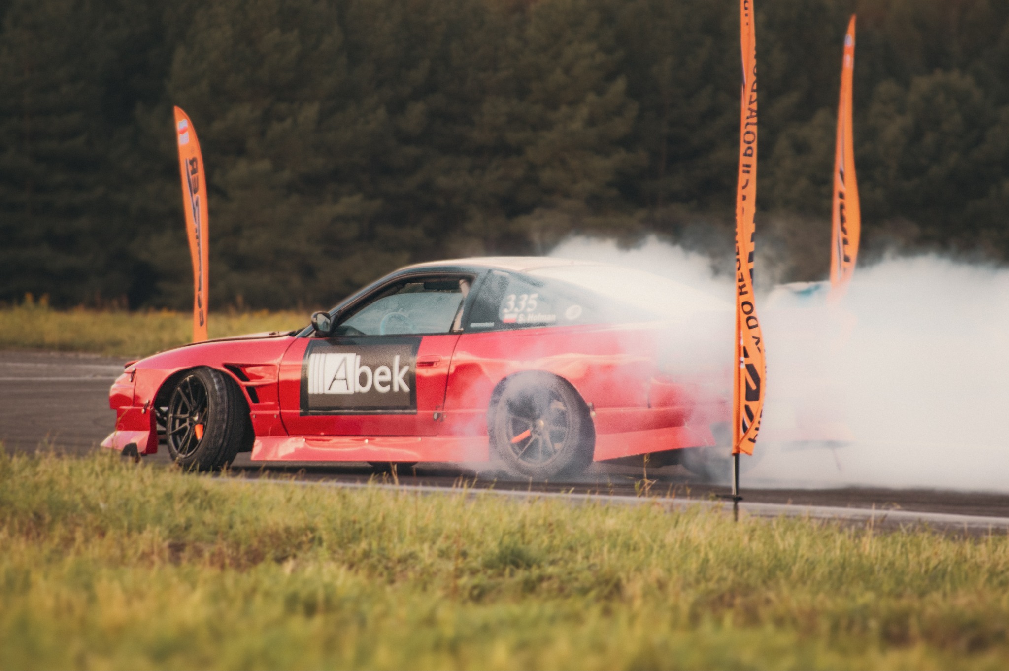 Czerwony samochód driftowy z naklejką Abek na boku, unoszący się w dymie na torze, z pomarańczowymi flagami w tle.
